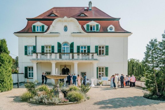 Helles, elegantes Gebäude mit grünen Fensterläden und einer festlichen Versammlung im Garten.