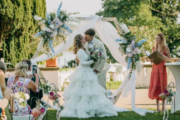 Brautpaar beim Hochzeitsempfang unter einem Blumenbogen, mit Gästen im Hintergrund.